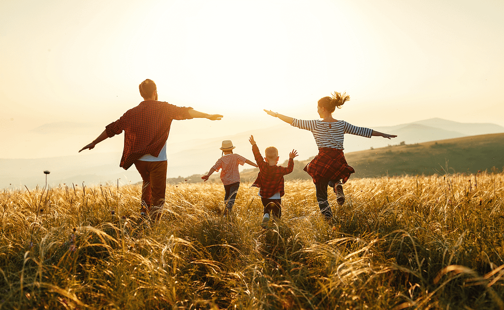 A imagem captura uma família de quatro pessoas caminhando em um campo de grama alta sob a luz quente do sol poente ou nascente. Os pais estão à frente, de costas para a câmera e de braços abertos, como se estivessem abraçando a natureza ou celebrando um momento de liberdade. Os dois filhos, um pouco atrás dos pais, também estão com os braços levantados, expressando alegria e energia enquanto correm pelo campo. Ao fundo, suaves colinas se estendem sob um céu claro com tons de amarelo e branco do sol. A atmosfera geral é de felicidade, união familiar e conexão com a natureza.