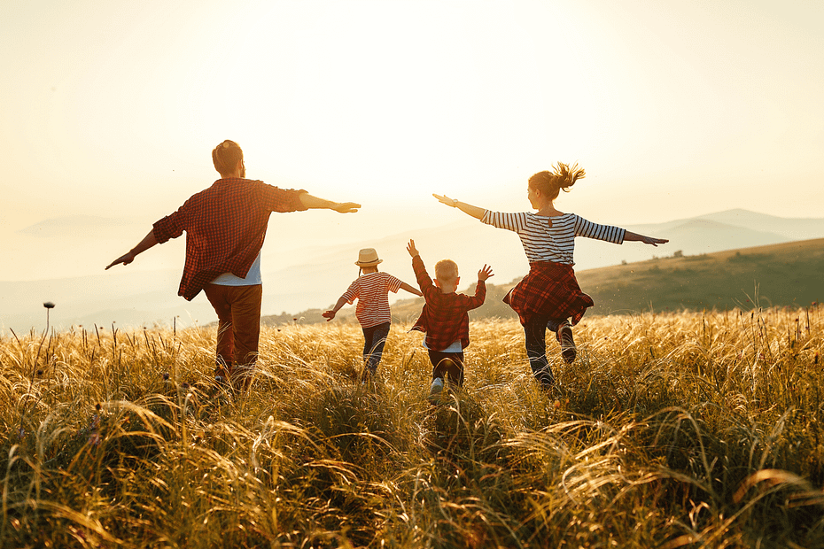 A imagem captura uma família de quatro pessoas caminhando em um campo de grama alta sob a luz quente do sol poente ou nascente. Os pais estão à frente, de costas para a câmera e de braços abertos, como se estivessem abraçando a natureza ou celebrando um momento de liberdade. Os dois filhos, um pouco atrás dos pais, também estão com os braços levantados, expressando alegria e energia enquanto correm pelo campo. Ao fundo, suaves colinas se estendem sob um céu claro com tons de amarelo e branco do sol. A atmosfera geral é de felicidade, união familiar e conexão com a natureza.